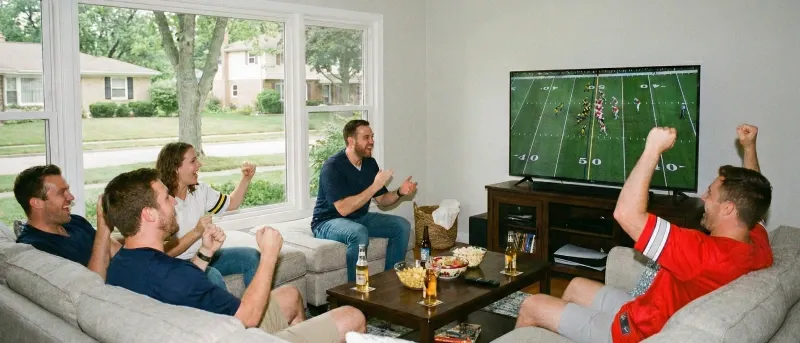 A group of excited friends cheer while watching a football game on TV in a living room.
